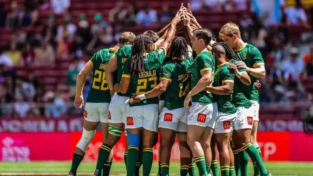 A South African rugby team huddles together on the field, raising their hands in unity before a match.