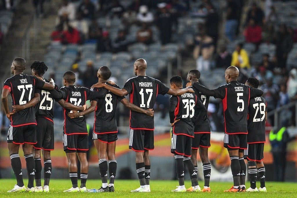Orlando Pirates players stand arm in arm, facing away in unity before a match.