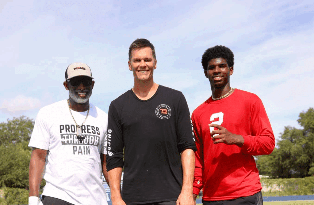 Tom Brady poses with two individuals, one in a "Progress Through Pain" t-shirt and the other wearing a red number 2 shirt, on a football field.